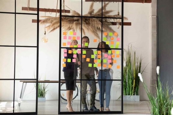 Two individuals stand in front of a glass board adorned with sticky notes, embodying a collaborative and inclusive meeting environment adaptable for neurodivergent staff