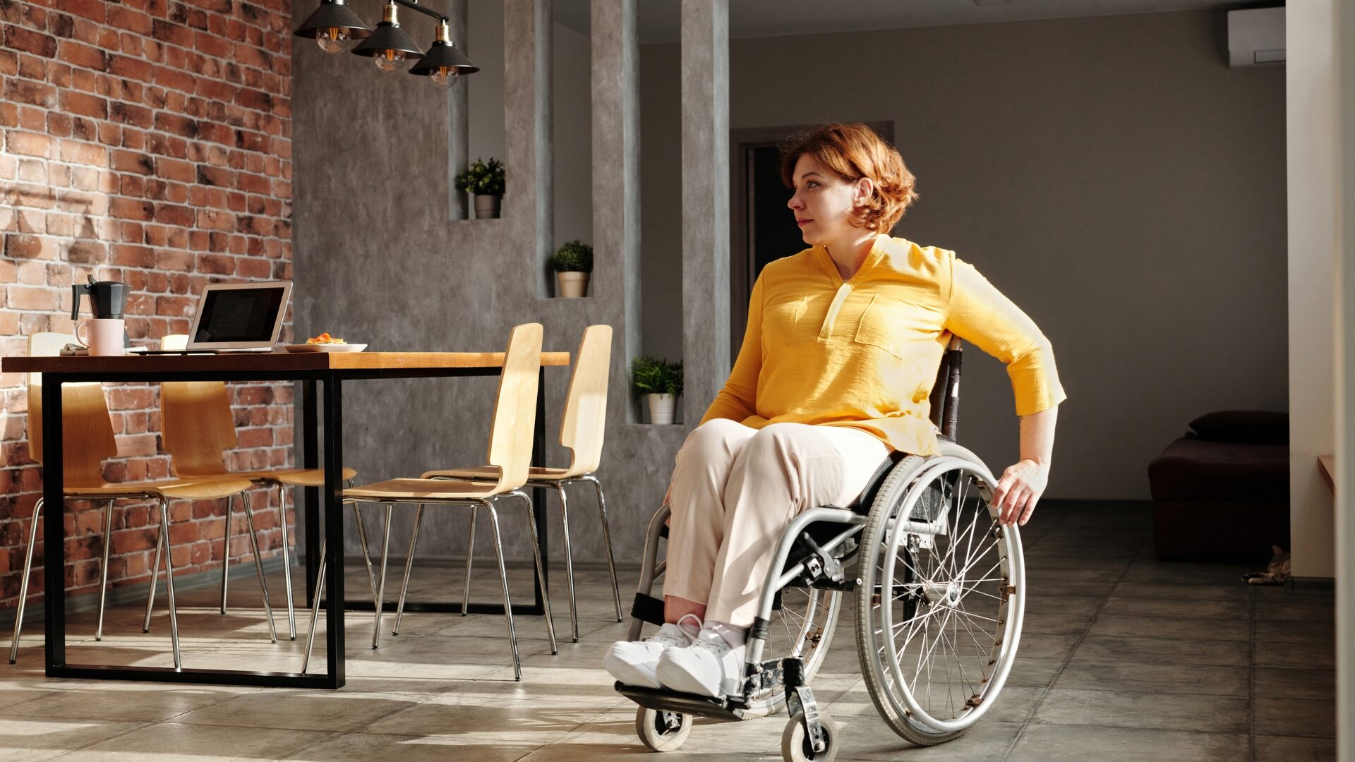 Woman in a wheelchair at a café table with a laptop, looking away thoughtfully.