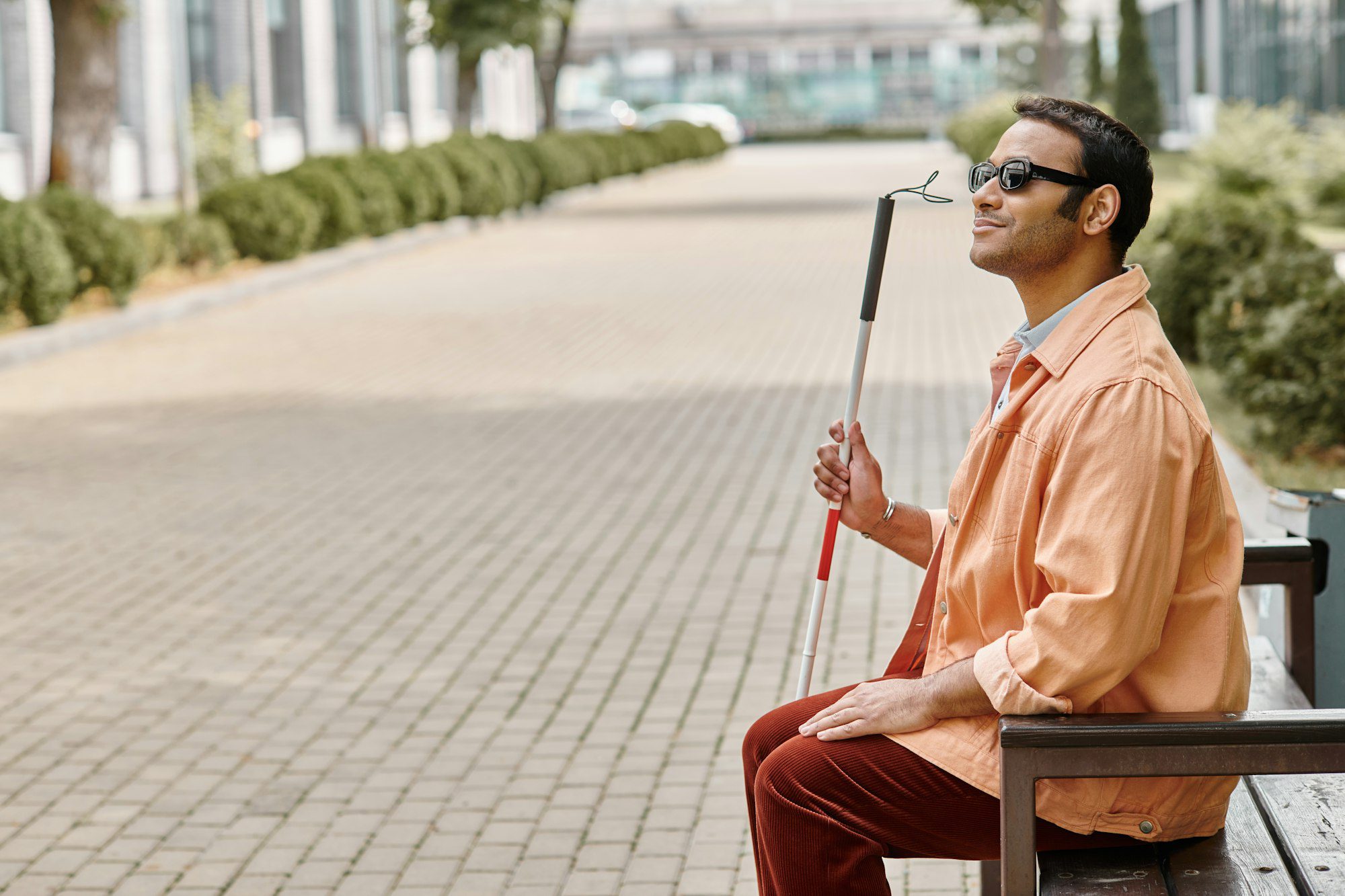 glad indian blind man in orange jacket sitting outside on bench with walking stick and glasses