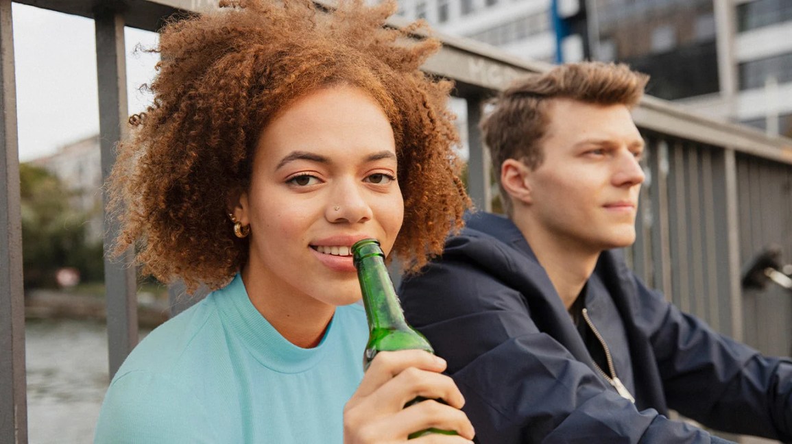 A woman drinking from a glass bottle while sitting next to a man.