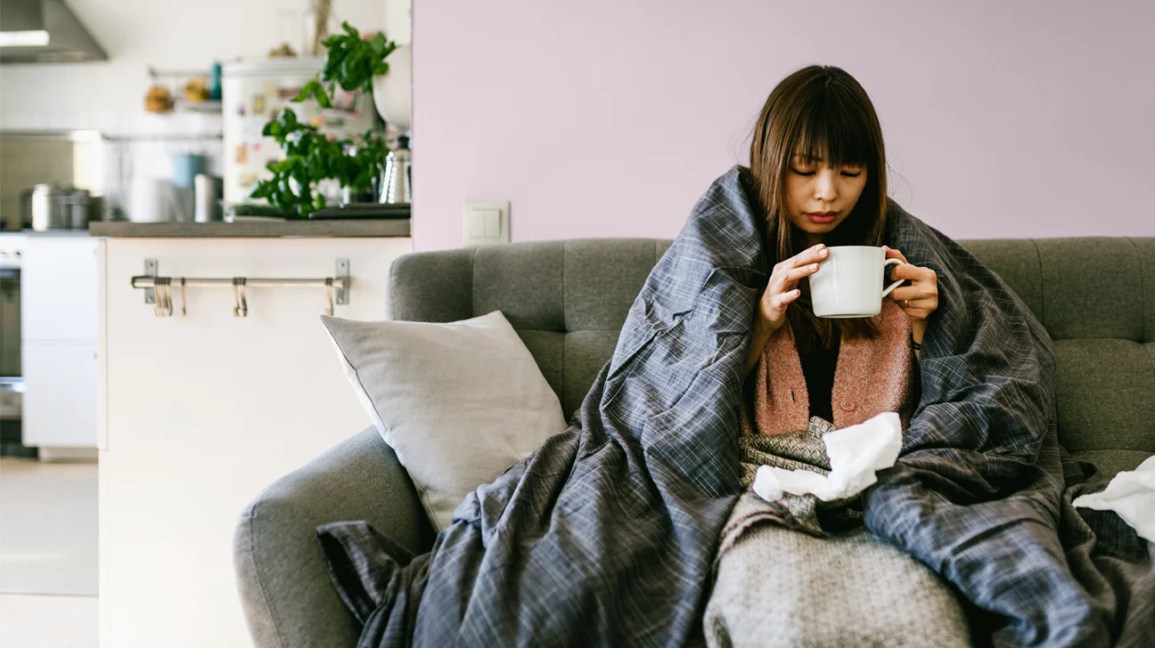 A woman sits on the couch feeling ill, showing symptoms that could be flu or covid-19. 