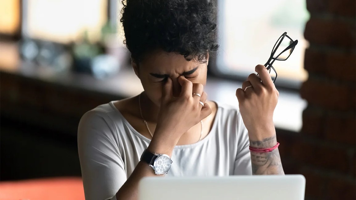 A woman sits in front of a laptop, pinching the bridge of her nose due to a headache. 