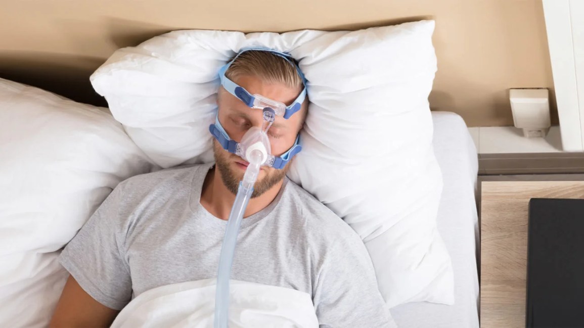 A man lies on his back in bed with a CPAP mask on his face. 