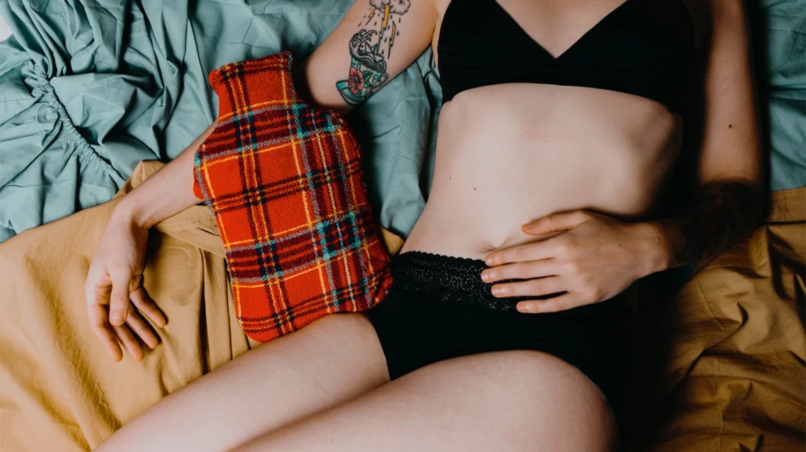 A woman lies on a bed wearing underwear, with her hand on her abdomen. 