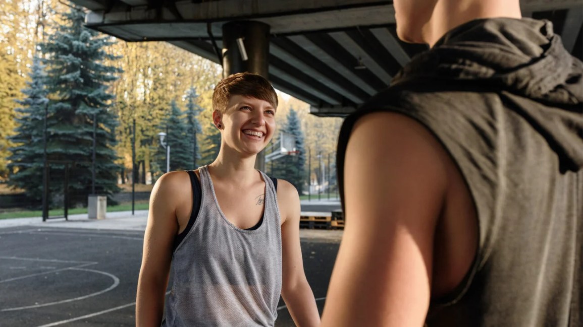 person with short hair and a visible shoulder tattoo smiling at the person they're playing basketball with