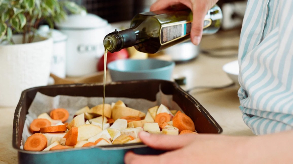 A person pours olive oil onto a baking sheet covered with food.