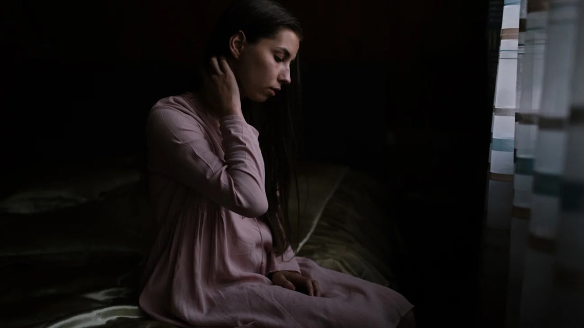 Person wearing pink nightdress sits on bed in dark looking distressed