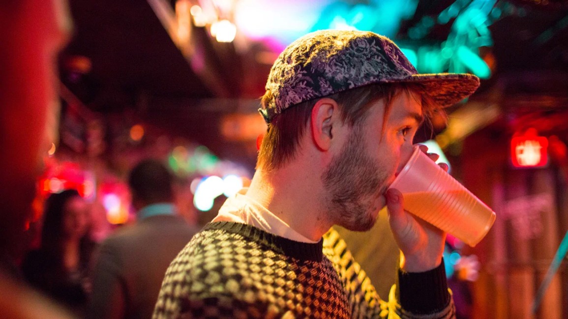 man in yellow checked aweater and baseball cap drinking beer out of cup 1