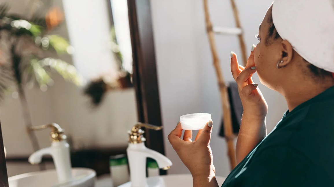 person applying moisturizer to face in front of a bathroom mirror