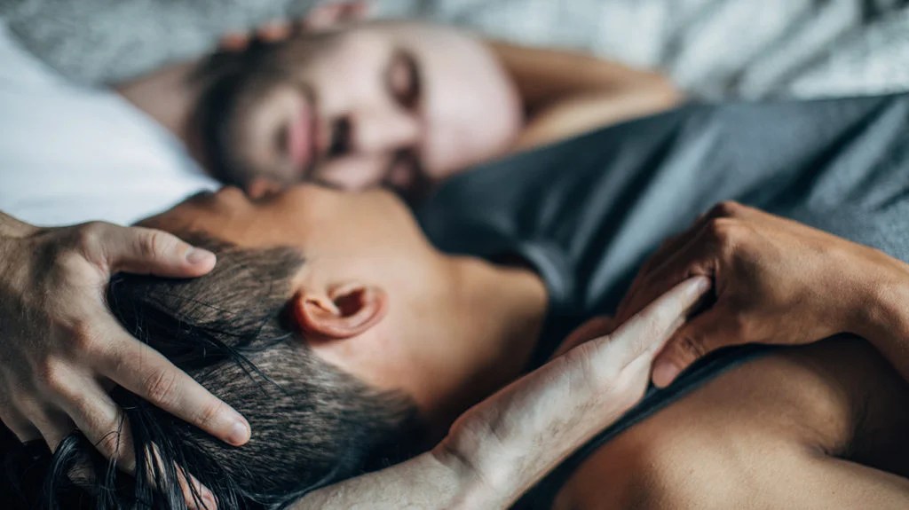 a homesexual couple lying in bed together discussing Can HSV2 be transmitted through oral sex?