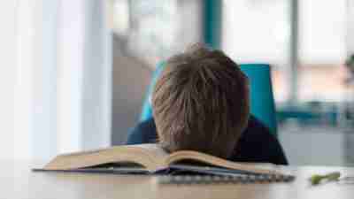 Tired 8 years old boy doing his homework at the table. Child reading a book at the desk.