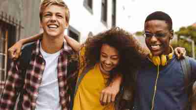 Close up of three college friends standing in the street with arms around each other. Cheerful boys and a girl wearing college bags having fun walking outdoors.