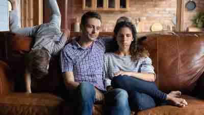 Exhausted young Caucasian parents sot on sofa at home annoyed by ill-behaved small kids play have fun. Unhappy upset mom and dad distressed tired by naughty active children. Upbringing concept. fizkes/Getty Images