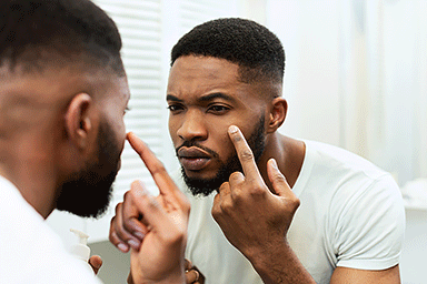 Young African American man touching face, examining quality skin, looking at mirror in bathroom