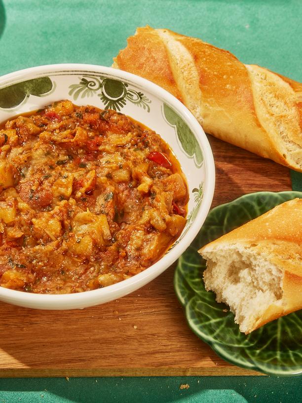 Zaalouk: "Displaying the completed Zaalouk recipe in a bowl, adorned with vibrant greens, placed on a wooden board alongside a plate of bread and a sliced baguette, set against a lush green backdrop."