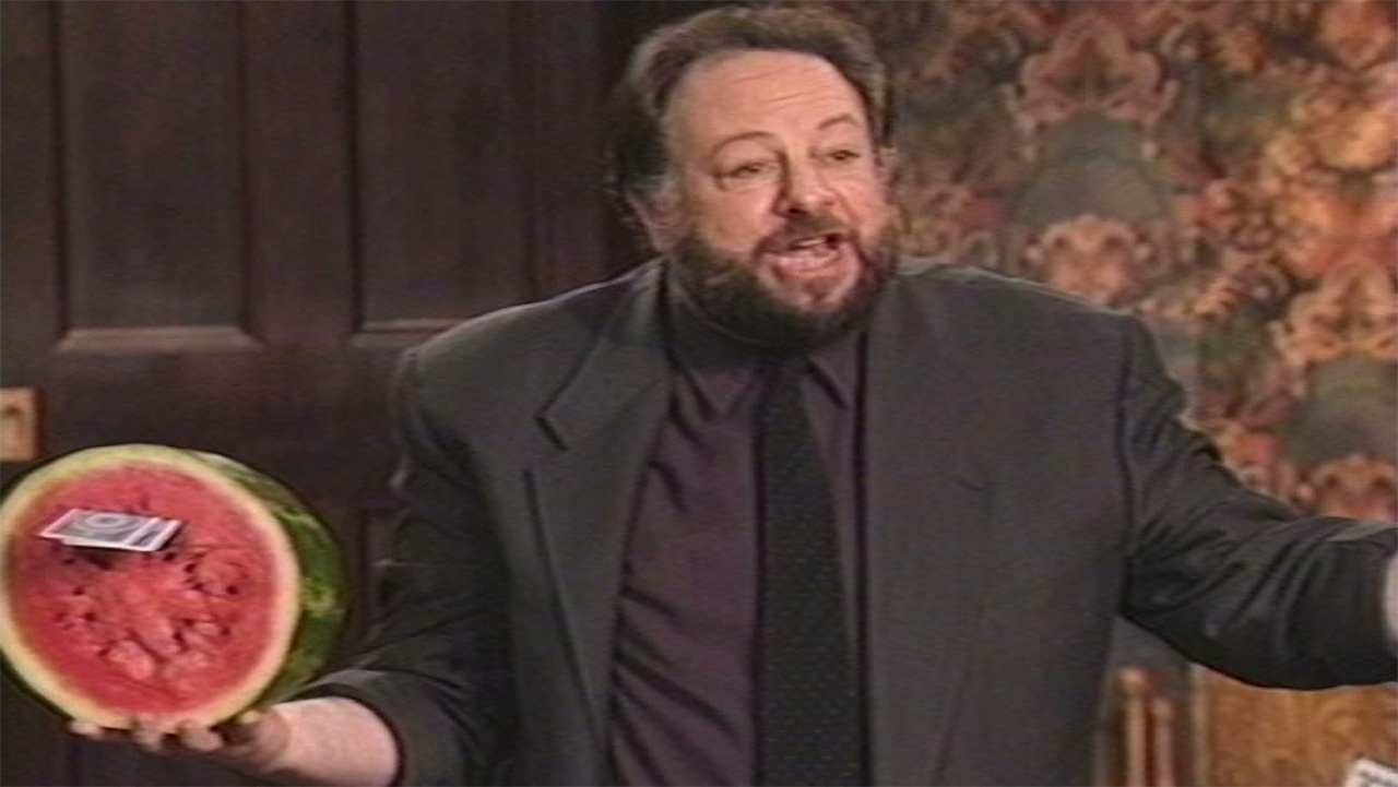 Ricky Jay, with his hair balding halfway up his head but black and tall, and a large black beard, in a black suit with a black tie and a dark purple shirt, looking at the camera holding a cut-in-half watermelon with two playing cards into the inside.