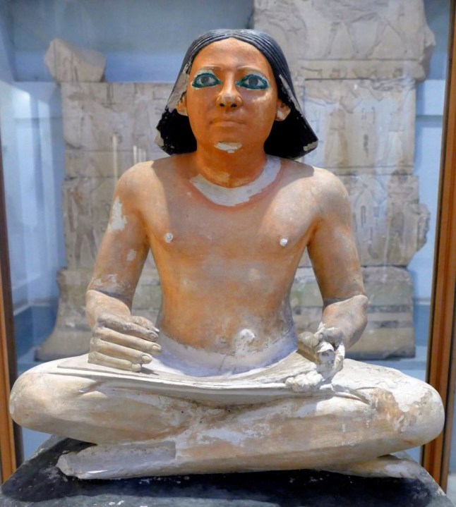 painted limestone statue of an Egyptian scribe seated cross-legged holding an open papyrus scroll