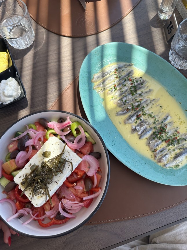 A Greek salad and a blue plate of anchovies on a restaurant table