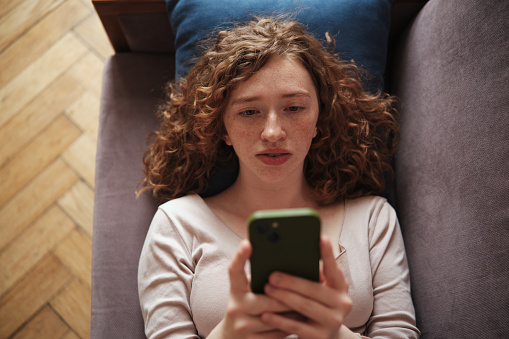 Sad lonely gen z girl lying on sofa with phone, scrolling social media, Internet addiction, wasting time. Mental health of Millennial generation people