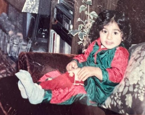 An old photo as Sharan as a toddler, sitting on her sofa, wearing a red and green dress