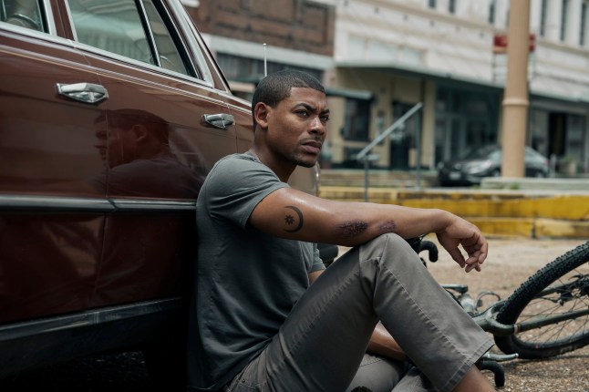 Aaron Pierre as Terry Richmond in a scene from Rebel Ridge, sat on the floor leaning against a brown car