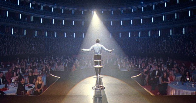 A scene from Robbie Williams biopic Better Man showing a man from the back standing onstage in a spotlight with an audience in front of him