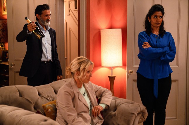 Nish holding up a bottle of champagne while Suki stands with her arms crossed and Kathy sits in an armchair in EastEnders