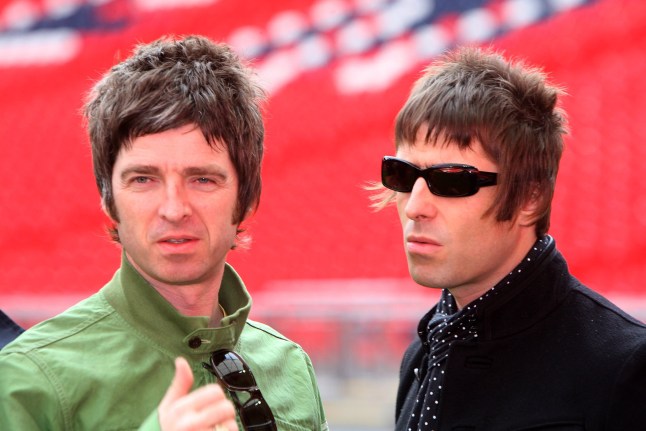 Noel and Liam Gallagher stood together at Wembley Stadium. Noel wears a green jacket while Liam wears all black and dark sunglasses