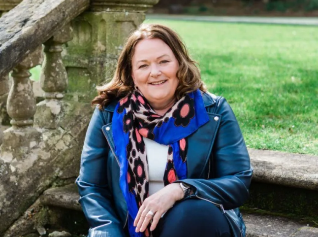 Alison sitting on steps in a large garden, wearing a navy jacket and colourful scarf