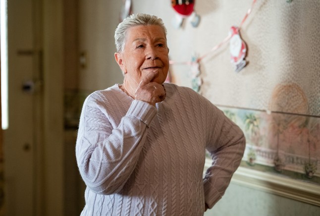 Mo stands grinning in the hallway in EastEnders