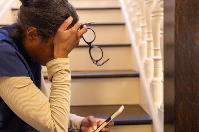 black woman wearing medical scrubs sitting on steps using smart phone with eyeglasses off & forehead in palm of hand