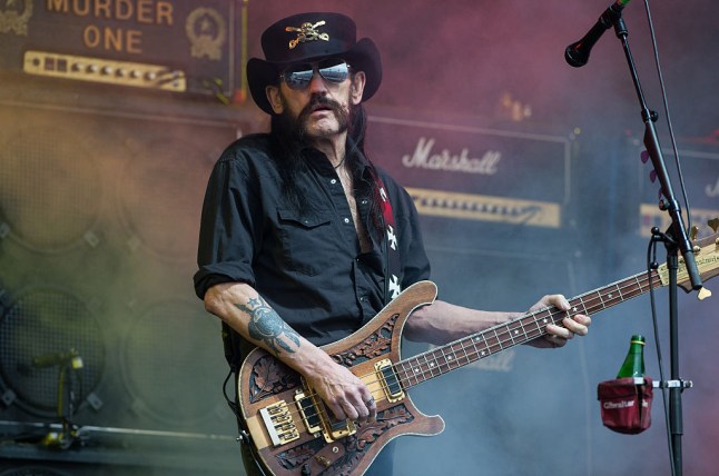 Lemmy on stage holding a brown guitar in front of a microphone stand, wearing a black shirt, sunglasses and large black hat