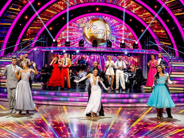 Several Strictly couples stand in the ballroom, surrounded by bright pink and yellow stage lights and with the gold Strictly glitterball behind them