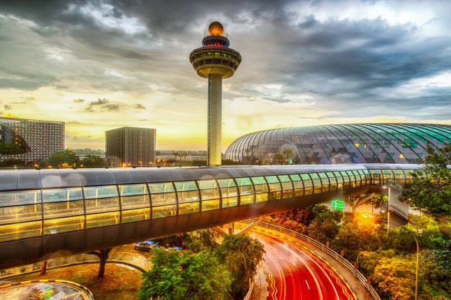 Exterior of Changi Airport's Terminal 2 in Singapore, showing a lit-up passenger transit tunnel and control tower at dusk