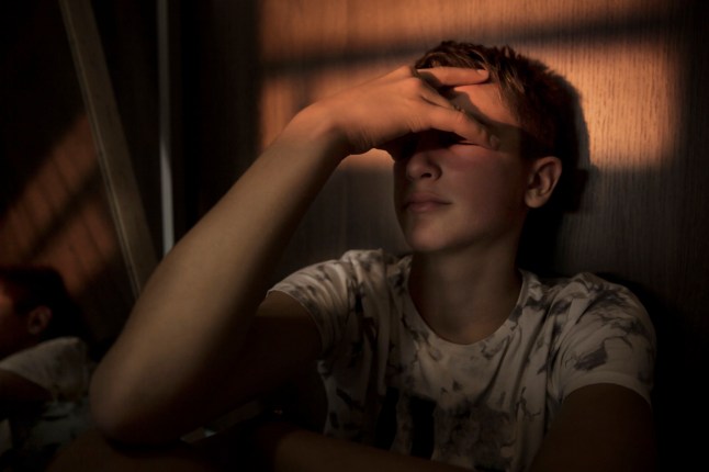 A teenager holding his face clearly upset and sad in a dark room.