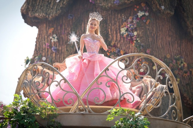 Ariana Grande as Glinda in a pink gown, holding a wand and wearing a crown while stood on a platform in Munchkinland, in a scene from the film Wicked