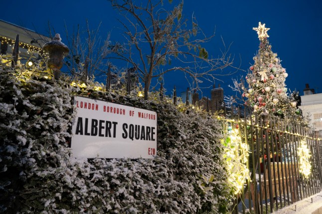 The Albert Square sign on the railings, covered in snow, with a Christmas tree in the background and Christmas lights around in EastEnders