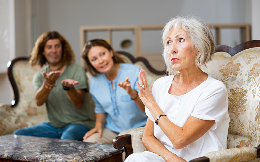 Angry mature female in armchair, keeping distance from the couple sitting on the couch at home