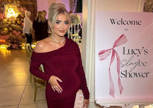 Lucy Fallon in a dark red, off-shoulder dress holding her baby bump in front of a sign that reads 'Welcome to Lucy's slaybe shower'