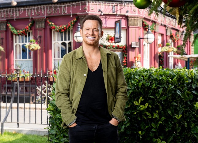 Joe Swash as Mickey Miller, smiling with his hands in his pockets in front of the Queen Vic in EastEnders