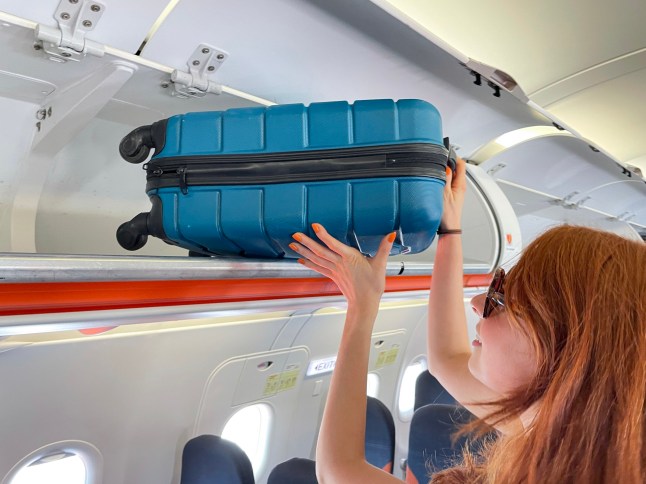 Stock photo showing close-up view of row of carryon luggage overhead baggage compartments in an airplane cabin with beautiful red haired woman putting away her suitcase.