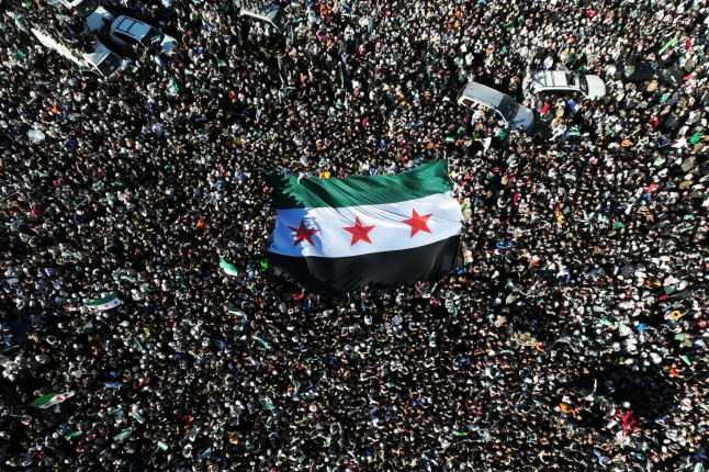 An areal photo shows crowds of Syrians raising a giant independence-era flag, used by the opposition since the uprising began in 2011, as they celebrate the fall of Bashar al-Assad earlier this week at the central Umayyad Square in Damascus on December 13, 2024. Thousands of jubilant Syrians also gathered outside a landmark mosque in the capital Damascus to celebrate during the first Friday prayers since Assad's ouster on December 8. (Photo by Omar HAJ KADOUR / AFP) (Photo by OMAR HAJ KADOUR/AFP via Getty Images)