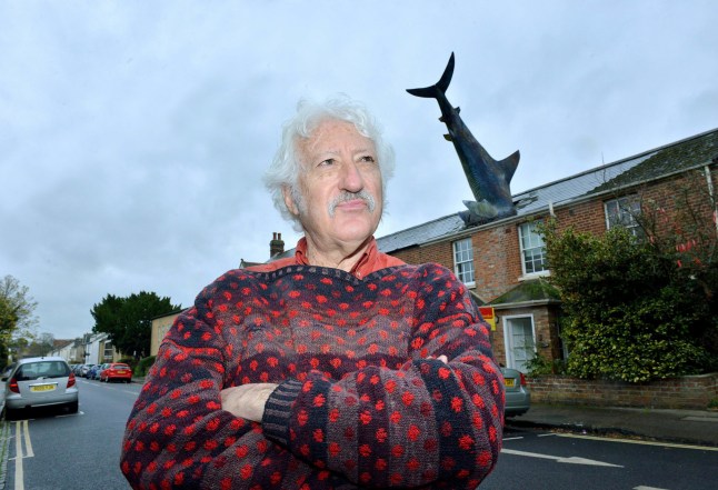 FILE PICTURE - Bill Heine outside his house number two New High Street in Oxford. Photo released December 16 2024. The owner of the iconic "shark house" in Oxford says "come while you can" after council demand closure of quirky Airbnb.Oxford City Council won the planning appeal banning the use of short let accommodation last week for the quirky property in New Street, Headington, more widely known as the Shark House - which features a 25ft shark sticking out of its roof.This comes after owner Dr. Magnus Hanson-Heine, whose father Bill Heine constructed the sculpture without official planning permission from Oxford City Council in 1986.For the last five years, Dr. Hanson-Heine has been letting the property out on Airbnb for short term stays - but received notice from the council to shut it down, after a member of the public complained about the change from a residential use to a short-term let.