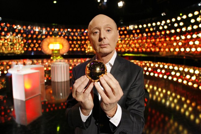 Jasper Carrott standing in the Golden Balls studio, holding a golden ball and looking quizzical.