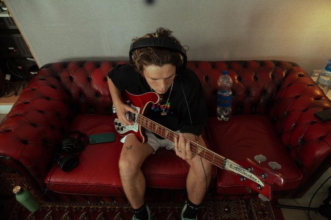 Cruz Beckham sitting on a red leather sofa in a T-shirt and shorts playing an electric guitar