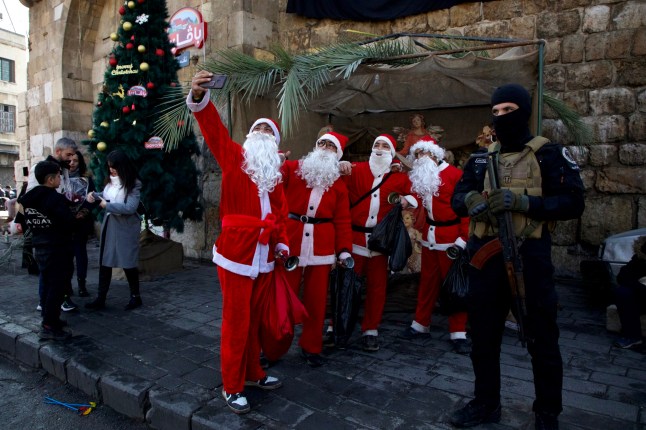 DAMASCUS, SYRIA - DECEMBER 31: People dressed as Santa Claus (Father Christmas) pose for a selfie while a man holding a weapon stands guard on New Year's Eve on December 31, 2024 in Damascus, Syria. Syria is transitioning after the fall of the Baath regime and the Assad family's rule, with a temporary administration now in place in Damascus and Aleppo. (Photo by Ali Haj Suleiman/Getty Images) *** BESTPIX ***