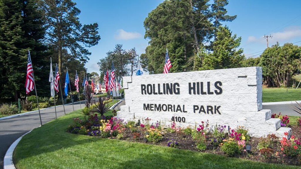 Marble sign reading "Rolling Hills Memorial Park" with flowers planted in front of it and American flags lining roadway entrance into cemetery.