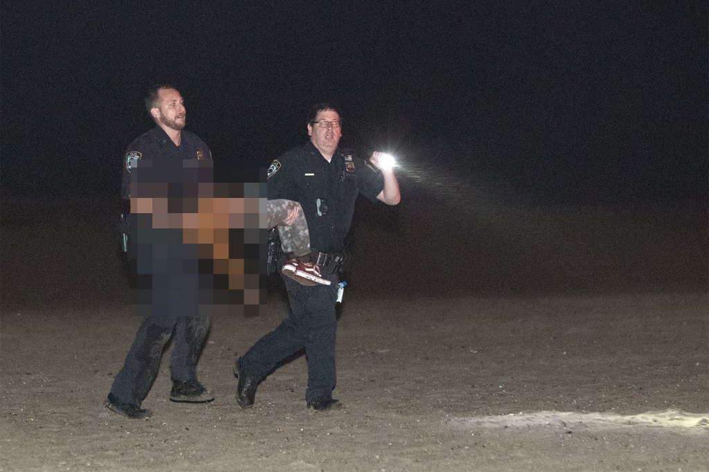 Two police officers carry one of Erin Merdy's children (whose body has been blurred) off the beach at night.