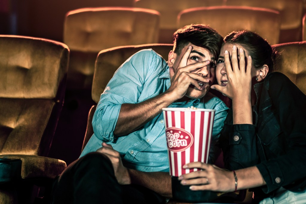 A shocked couple watching a movie in a theater, holding popcorn and closing their eyes