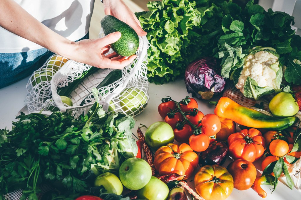 Young woman returned with purchases from grocery store takes fresh organic vegetables out of mesh bag putting on kitchen table at home close view